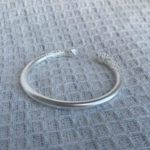 May include: A silver-toned open cuff bracelet featuring a smooth, rounded band that transitions into intricate, embossed scrollwork patterns at both ends. The metal has a matte finish, and the design terminates in rounded, decorative terminals.