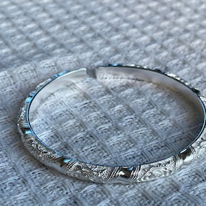 May include: A silver-toned open cuff bracelet featuring a repeating pattern of engraved floral motifs alternating with smooth, polished vertical bands. The metal has a reflective finish and a circular shape with a small gap at the top for adjustment.