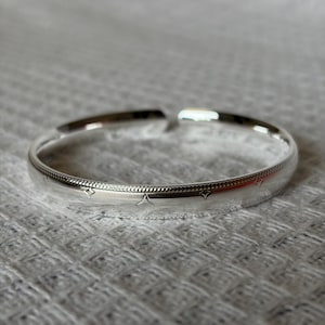 May include: A polished silver-tone open cuff bracelet featuring a textured band with a delicate beaded edge and small engraved star-shaped motifs spaced evenly along the circumference of the metal surface.