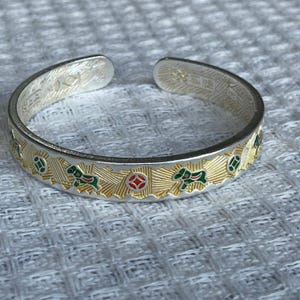 May include: A silver-toned open cuff bracelet featuring a textured gold-colored band with repeating enamel motifs of green horses and red-centered circular coins. The interior of the band displays engraved Chinese characters and a textured finish.