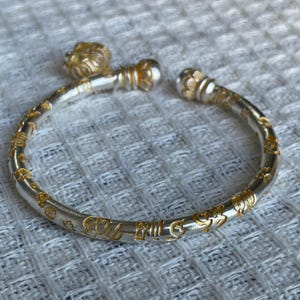 May include: A silver-toned open cuff bracelet featuring a matte finish with gold-colored embossed symbols and characters along the band. The bracelet ends are capped with rounded, textured decorative elements, one of which is shaped like a small head.