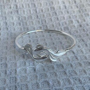 May include: A polished silver-toned bangle bracelet featuring a central decorative element shaped like a stylized fish with flowing fins. The metal surface is smooth and reflective, showcasing intricate detailing on the fins and body of the fish.