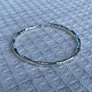 May include: A silver-toned open cuff bracelet featuring a segmented design that mimics the appearance of bamboo stalks. The metal has a polished, reflective finish and the circular band is interrupted by a small gap at the top of the structure.