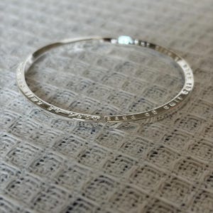 May include: A silver-toned open cuff bracelet featuring a thin, circular band with a polished metallic finish. The outer surface of the bracelet is engraved with small, legible text that runs continuously along the entire length of the band.