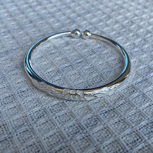 May include: A polished silver cuff bracelet featuring an open-ended design with two small spherical terminals. The outer surface of the metal band is decorated with an intricate, engraved floral pattern that spans the front section of the jewelry.
