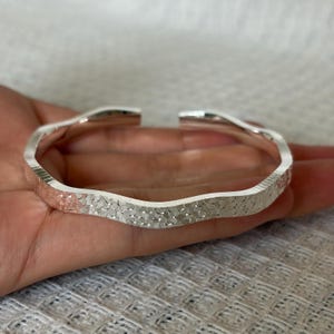 May include: A silver-colored open cuff bracelet featuring a wavy, undulating band design. The surface of the metal has a textured, hammered finish that reflects light across its entire length, creating a shimmering appearance on the exterior.