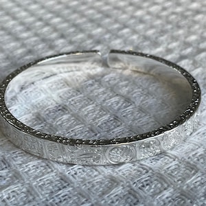 May include: A silver-colored open cuff bracelet featuring a repeating embossed pattern of circular and rectangular motifs along the entire outer surface. The metal has a polished, reflective finish with a distinct gap at the top of the band.