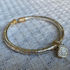 May include: A silver-toned open bangle bracelet featuring a matte finish and gold-colored embossed symbols along the band. A small, dangling charm shaped like a beckoning cat with gold-colored accents is attached to one end of the circular bracelet.