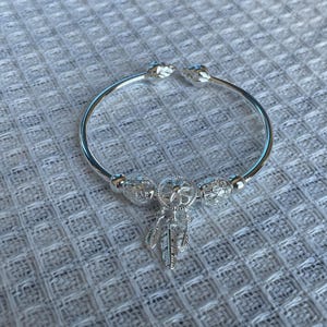 May include: A silver-toned open cuff bracelet featuring a central circular charm with a webbed pattern and three dangling feather-shaped charms. The bracelet is accented with two spherical beads with intricate cut-out designs on either side.
