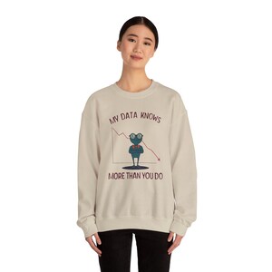 Puede incluir: Una sudadera beige de cuello redondo con un gráfico de un alienígena azul con gafas y pajarita roja. Detrás hay un gráfico de líneas rojas descendentes. El texto dice MY DATA KNOWS MORE THAN YOU DO en letras mayúsculas de color rojo oscuro.