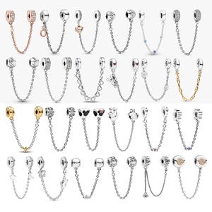 May include: A collection of twenty-four silver, gold, and rose-gold tone safety chains for charm bracelets. Each piece features two cylindrical beads connected by a delicate metal chain, with various decorative motifs including hearts, stars, and stones.