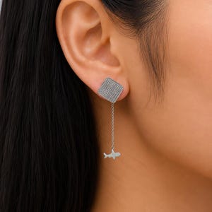 May include: A silver-toned earring featuring a square stud with a textured, cross-hatched surface. A delicate silver chain hangs from the bottom of the square, ending in a small, flat, silver-toned shark-shaped charm that dangles freely.