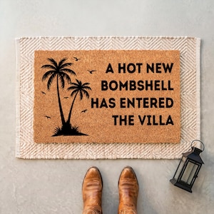 May include: A rectangular tan coir doormat featuring a black graphic of two palm trees and several birds. The text A HOT NEW BOMBSHELL HAS ENTERED THE VILLA is printed in bold black capital letters on the right side of the mat surface.