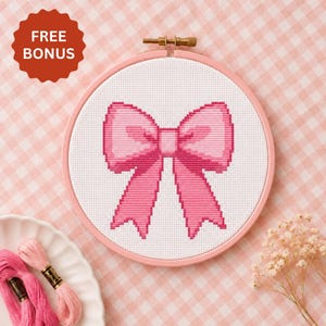 May include: A cross-stitch embroidery project featuring a pink bow on white fabric, secured in a round pink hoop. The design includes a red scalloped badge with the text FREE BONUS. Two skeins of pink embroidery floss are visible nearby.