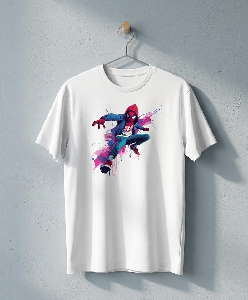 May include: A white short-sleeved t-shirt featuring a graphic print of a character wearing a red hooded jacket, blue pants, and red shoes, depicted in a dynamic pose with abstract blue and pink watercolor-style paint splatters surrounding them.