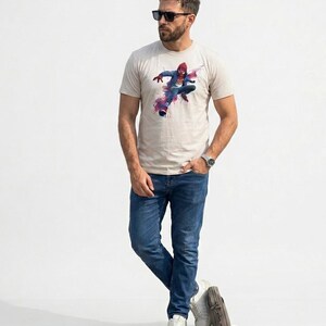 May include: A beige short-sleeved t-shirt featuring a graphic print of a superhero in a red and blue suit leaping forward with pink and purple watercolor-style splashes. The shirt is paired with blue denim jeans, white sneakers, and sunglasses.