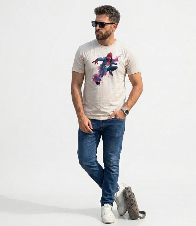 May include: A beige short-sleeved t-shirt featuring a graphic print of a superhero in a red and blue suit leaping forward with pink and purple watercolor-style splashes. The shirt is paired with blue denim jeans, white sneakers, and sunglasses.