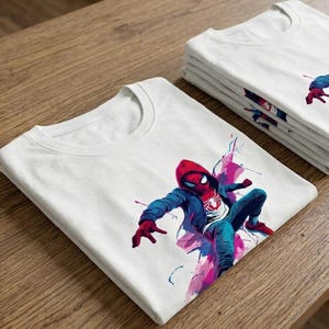 May include: A white short-sleeved t-shirt featuring a graphic print of a character wearing a red mask, a blue hooded jacket, and blue pants, depicted in a dynamic pose with vibrant pink and blue watercolor-style paint splatters around the figure.