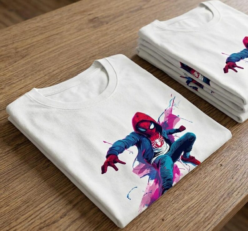 May include: A white short-sleeved t-shirt featuring a graphic print of a character wearing a red mask, a blue hooded jacket, and blue pants, depicted in a dynamic pose with vibrant pink and blue watercolor-style paint splatters around the figure.