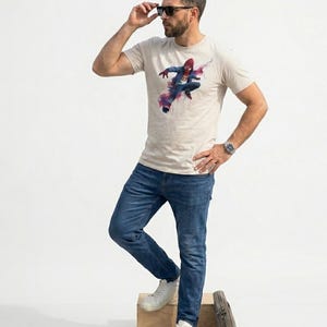 May include: A beige short-sleeved crew neck t-shirt featuring a graphic print of a superhero in a red and blue suit mid-action, accented with abstract pink and purple paint splatters. The shirt is paired with blue denim jeans and white sneakers.