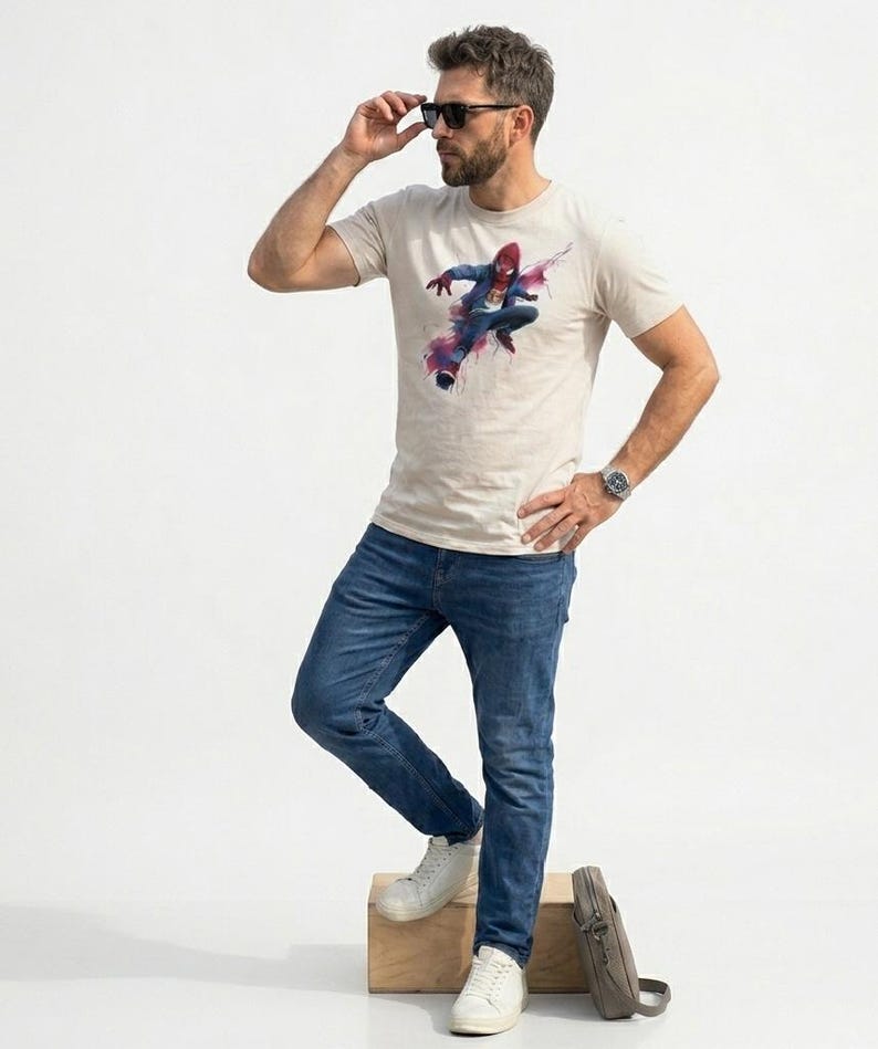 May include: A beige short-sleeved crew neck t-shirt featuring a graphic print of a superhero in a red and blue suit mid-action, accented with abstract pink and purple paint splatters. The shirt is paired with blue denim jeans and white sneakers.