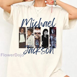 May include: A cream-colored short-sleeve t-shirt featuring a graphic print of Michael Jackson. The design includes the name Michael Jackson in dark blue script text and five vertical panels showing various images of the artist.