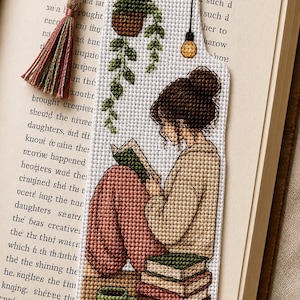 Peut inclure: Un marque-page rectangulaire en point de croix représentant une personne avec un chignon brun lisant un livre, assise près d'une pile de livres et d'une tasse verte. Au-dessus, une plante suspendue et une ampoule sont brodées avec un pompon.