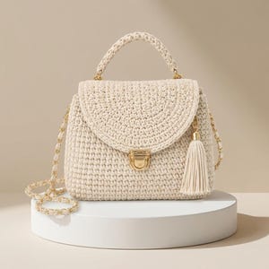 May include: A cream-colored crochet handbag featuring a structured top handle and a matching shoulder strap woven with gold-tone chain links. The bag has a curved flap closure secured with a gold-tone metal clasp and a decorative matching tassel.