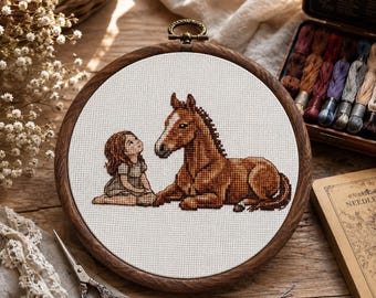 May include: A cross-stitch embroidery piece in a circular wooden hoop featuring a brown horse lying down next to a small child with brown hair. The design is stitched onto white fabric, showcasing detailed shading in the horse's coat and hair.