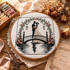 May include: A circular wooden embroidery hoop containing a cross-stitch pattern of a couple standing on a bridge under a floral arch. The design features black silhouettes of the couple, a black bridge, hanging lanterns, and pink roses with green leaves.