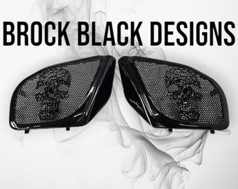 May include: A pair of black speaker grills for 23.5+ Road Glide motorcycles, featuring a glossy finish and a mesh design with a prominent skull motif in the center of each. The text BROCK BLACK DESIGNS is displayed above the speaker grills.