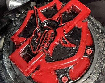 Puede incluir: Una tapa de depósito de combustible para motocicleta en rojo y negro brillante con un emblema en relieve de una criatura con cuernos. El accesorio circular está montado sobre una superficie oscura y texturizada con pernos rojos.