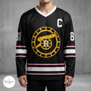Op de afbeelding: Een zwart ijshockey-shirt met een witte V-hals. Op de borst staat een geel cirkelvormig logo met een kanon en de letter R. De letter C staat linksboven op de borst. De mouwen en de zoom hebben witte en rode strepen met het nummer 81.