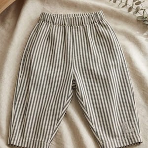 May include: A pair of pants featuring a vertical pinstripe pattern in dark gray and off-white. The pants have an elasticized waistband and a straight-leg cut, with a small, subtle embroidered logo detail visible on the lower left leg.
