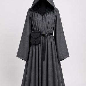 Pode incluir: Uma túnica com capuz cinza-carvão com mangas longas e largas e cintura franzida. A peça é ajustada com um cinto preto que segura uma pequena bolsa preta retangular com fecho de aba e uma fivela central na parte da frente.