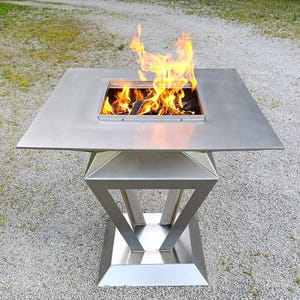Könnte beinhalten: Eine quadratische Feuerstelle aus Edelstahl mit einer flachen Oberseite und einer zentralen Öffnung, in der eine helle orangefarbene und gelbe Flamme brennt. Der Sockel hat ein geometrisches, kantiges Design, das sich zur Mitte hin verjüngt.