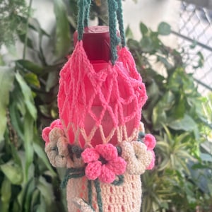 May include: A pink plastic bottle held in a handmade crochet holder. The holder features a pink net-style upper section, a cream-colored base, and decorative crochet flowers in shades of pink and cream with dark green stems and leaves.