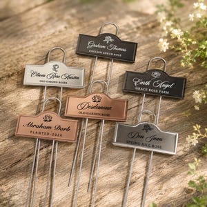 May include: Six metal garden plant markers with wire stakes, featuring rectangular plates in black, silver, and copper finishes. Each plate is engraved with a rose icon and text including names like Graham Thomas, Olivia Rose Austin, and Earth Angel.