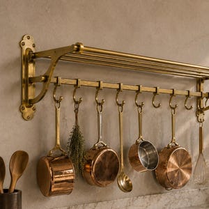 Op de afbeelding: Een aan de muur gemonteerd koperkleurig keukenrek met een bovenplank en een horizontale stang met meerdere S-haken. Aan de haken hangen diverse koperen pannen, een metalen garde, een metalen soeplepel en een bosje gedroogde kruiden.