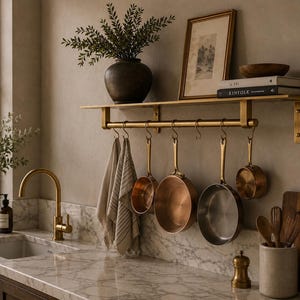 May include: A brass wall-mounted kitchen shelf with a hanging rail and six S-hooks. Four copper-toned pans of varying sizes are suspended from the hooks alongside two beige striped kitchen towels. The shelf holds a dark vase, a framed print, and books.