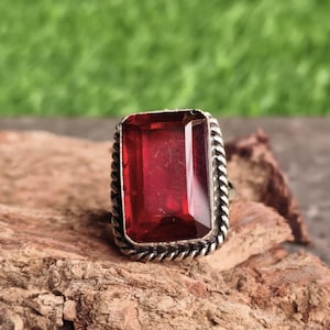 May include: A ring featuring a large, rectangular, faceted deep red gemstone set in a silver-toned metal frame. The setting is surrounded by a decorative twisted rope-like border, providing a textured edge to the central stone.