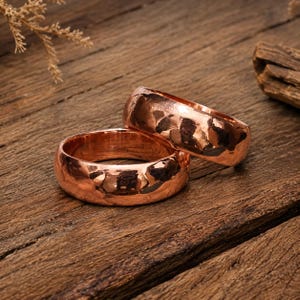 Puede incluir: Dos anillos de cobre pulido con una textura martillada distintiva. Los anillos presentan un perfil redondeado y un acabado metálico de color marrón rojizo cálido. La superficie del metal refleja la luz, resaltando las hendiduras irregulares.