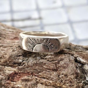 Puede incluir: Un anillo de metal plateado con un panel frontal plano y rectangular grabado con un diseño de rayos de sol. El motivo solar consiste en una base de semicírculo con líneas radiantes que se extienden hacia arriba, creando un patrón gráfico.
