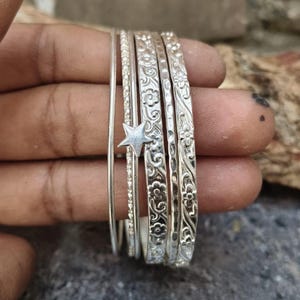 May include: A set of five silver-toned bangles featuring varied textures and designs. One bangle is smooth, one has a beaded texture, one features a small five-pointed star charm, and two are decorated with intricate floral and scroll patterns.