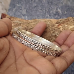 Pode incluir: Dois braceletes de metal prateado com padrões em relevo intrincados. Um bracelete exibe um motivo de folhas repetitivas, enquanto o outro apresenta uma série de pequenos detalhes circulares. Ambos têm um acabamento polido.