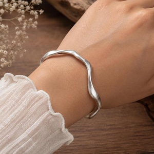 Puede incluir: Una pulsera rígida de tono plateado con un diseño liso y ondulado. La superficie metálica tiene un acabado pulido que refleja la luz a lo largo de su forma curva e irregular, creando una silueta continua y fluida alrededor de la muñeca.