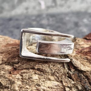 Puede incluir: Un anillo de tono plateado con un diseño geométrico de extremos abiertos y acabado liso y pulido. La banda consta de dos barras horizontales paralelas conectadas por un segmento vertical, creando una estructura de marco rectangular hueca.
