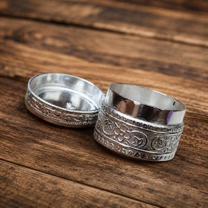 May include: A small, round, silver-coloured metal container with a separate lid. The exterior of both the base and the lid features a raised, embossed floral pattern with intricate scrolling details and a textured border along the top and bottom edges.