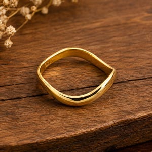 Può includere: Un anello in oro lucido con un design a fascia ondulata o sinuosa continua. La superficie metallica è riflettente e di colore uniforme, mostrando una forma semplice e curva che crea una silhouette quadrata morbida e arrotondata.