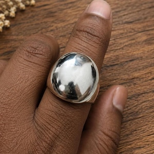 Puede incluir: Un anillo de tono plateado pulido que presenta una pieza central grande, lisa y redondeada en forma de cúpula. La superficie metálica es altamente reflectante, creando un acabado tipo espejo que capta la luz. La banda es gruesa y se estrecha.